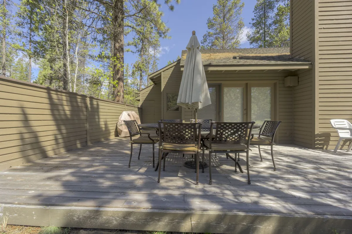 A serene patio with an outdoor dining table beneath a large umbrella, surrounded by mature trees. The scene conveys an atmosphere ideal for alfresco meals, morning coffee, or gathering with friends and family outdoors.