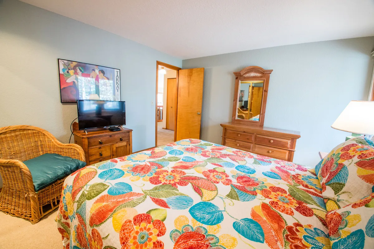 This image showcases a bedroom featuring colorful bedding, a wicker chair, a flat-screen television, and a wooden dresser. The space feels cozy and inviting, ideal for relaxing, watching TV, or reading a book, and hints at serene mornings or cozy movie nights.