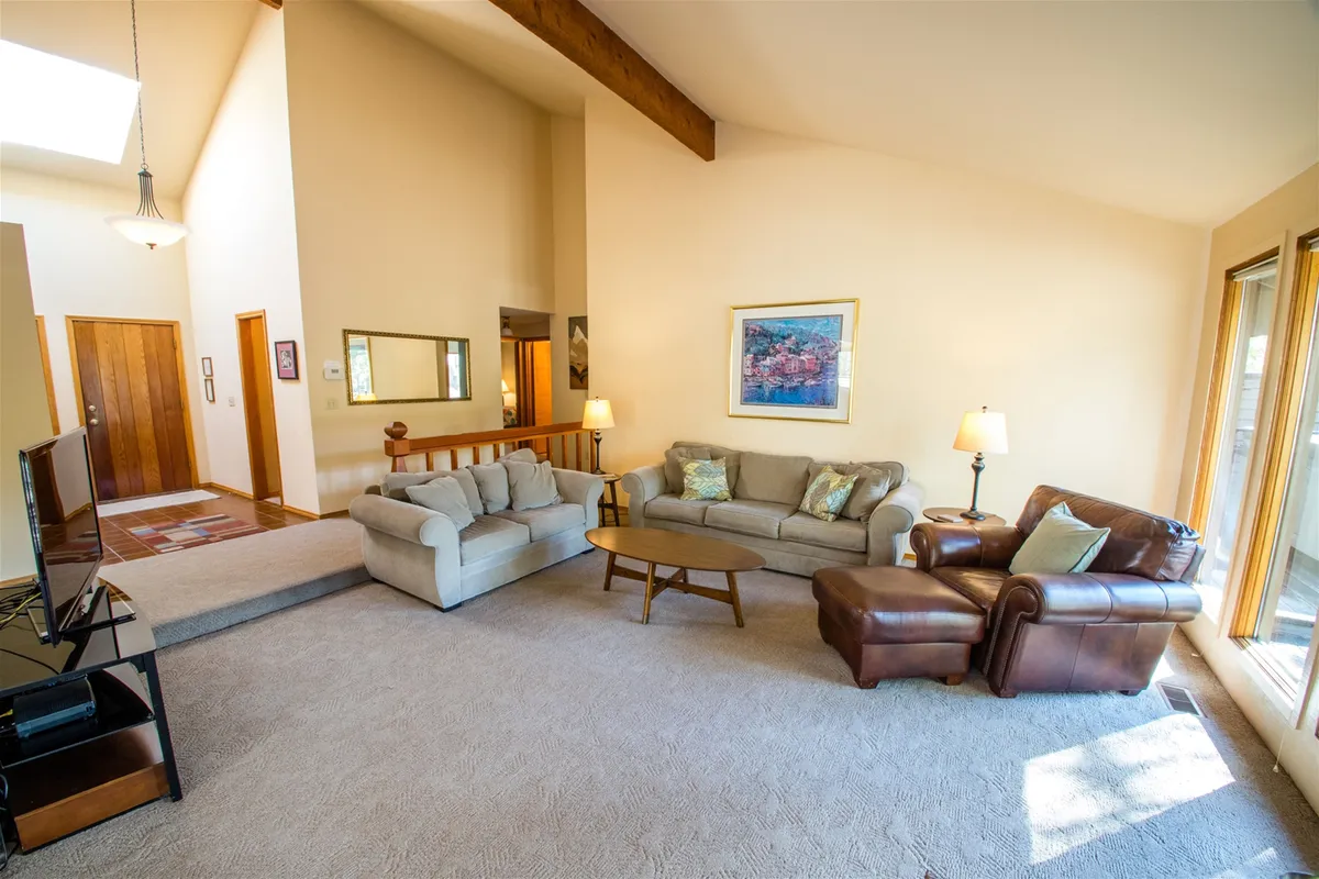 A spacious, comfortable living room with sofas, a coffee table, and large windows that offer glimpses of the wooded surroundings beyond. The space feels open yet cozy, perfect for relaxing with pets after a day outdoors.