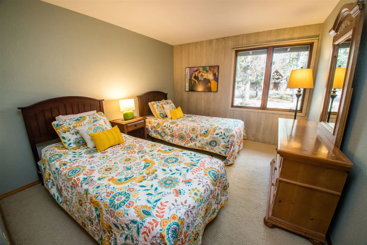 This inviting bedroom scene features two beds with vibrant quilts, plenty of natural light from a window, and a wooden dresser with a mirror. The room is designed for comfort and flexibility, ideal for accommodating a variety of group sleeping arrangements.