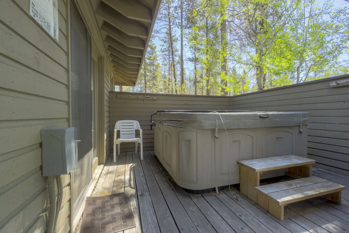 An exterior space featuring a private hot tub with wooden steps leading up to it and a patio chair nearby, all set beside the house and surrounded by trees. The area invites relaxation and immersion in nature, capturing the essence of a backyard retreat.