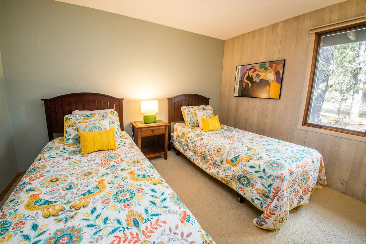 A cheerful bedroom with two twin beds adorned with colorful bedding, a nightstand with a lamp, ample natural light from the window, and decorative artwork on the wall. The space feels bright and flexible, perfect for families or friends.