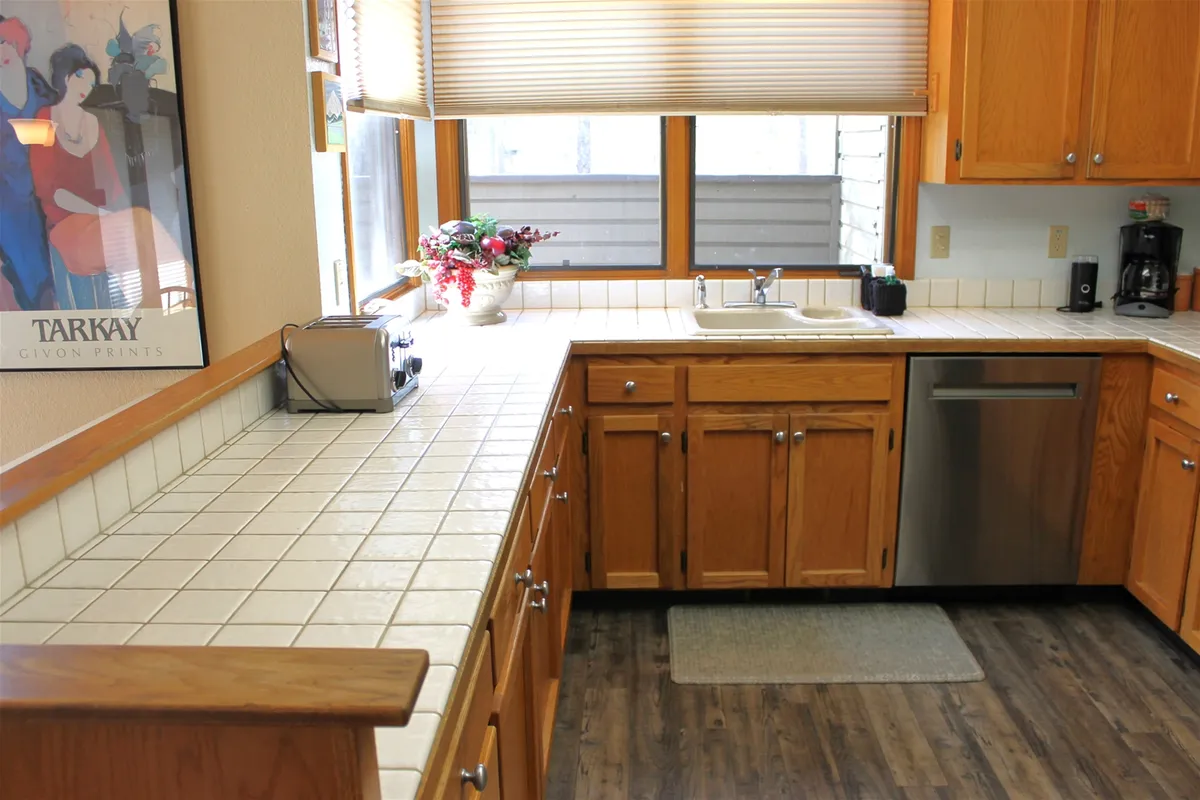 This image shows a kitchen area centered around a sink, set below a window that lets in abundant natural light. Wooden cabinetry, a generous countertop, and glimpses of kitchen appliances establish a welcoming and practical environment for meal prep and gatherings.